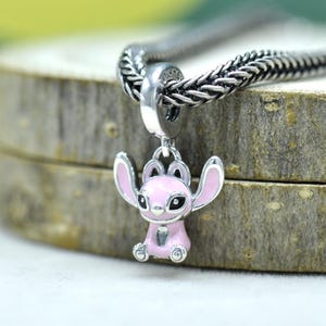 May include: A silver charm featuring a pink cartoon character with large ears and black eyes. The charm is suspended from a silver chain. The charm is a pendant, likely for a bracelet or necklace.