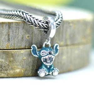 May include: A silver charm bracelet with a blue and silver Stitch charm. The charm is a cartoon character with large ears, a wide grin, and small feet. The charm is attached to a silver bracelet.