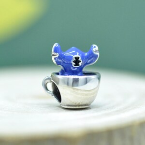 May include: A silver-toned metal charm bead shaped like a small teacup with a handle. Inside the cup sits a blue enameled character figure with large ears and a black nose detail. The charm features a hollow opening on the side for a bracelet chain.