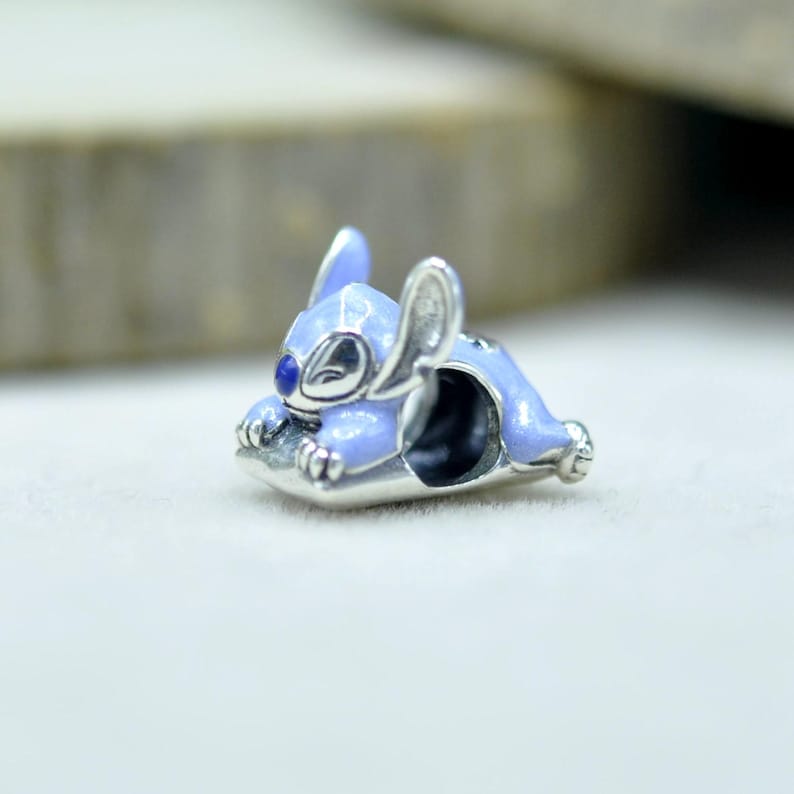 May include: A silver charm bead shaped like the character Stitch, featuring light blue enamel detailing on the body, large ears, and a dark blue nose. The charm has a hollow center for threading onto a bracelet, with polished silver accents.