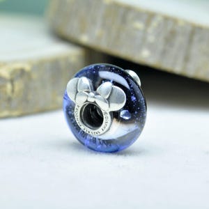 May include: A round, blue and silver charm with a Minnie Mouse bow design. The charm has a dark blue background with white speckles, and the bow is silver. The charm is marked with "PANDORA" and "&copy;Disney".