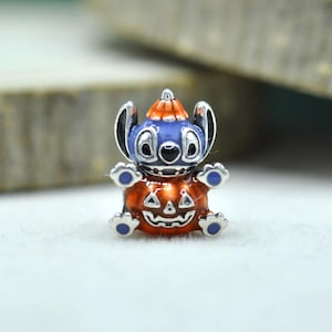 以下が含まれることがあります： オレンジ色のカボチャの帽子をかぶり、彫刻されたオレンジ色のジャック・オー・ランタンを持つ青いエイリアンのキャラクターが描かれたシルバーチャームビーズ。耳、足、カボチャの細部に青いエナメルのアクセントが施された光沢のある仕上げ。