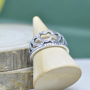 May include: A silver ring with a heart-shaped design. The ring features multiple heart cutouts and is adorned with small, sparkling accents. The ring is displayed on a wooden stand against a soft green background.