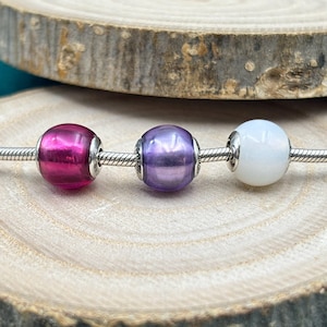 Può includere: Tre perline di vetro su una catena d'argento. Le perline sono rosa, viola e bianche.