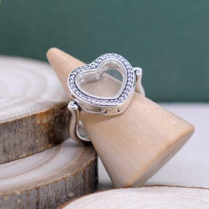Puede incluir: Un anillo plateado en forma de corazón, con un centro transparente y un borde de pequeñas piedras brillantes. El anillo se exhibe en un soporte de madera, mostrando su diseño y detalles.