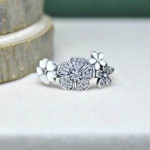 May include: A silver ring with a floral design. The ring features three flowers, the central one set with small, clear stones. The other two flowers are white with a single clear stone in the centre. The ring is displayed against a soft, neutral background.