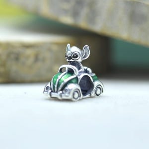 May include: A silver charm bead featuring the character Stitch driving a small green and silver car. The charm has a metallic finish with green enamel detailing on the hood and fenders, and a hollow center for a bracelet or necklace chain.