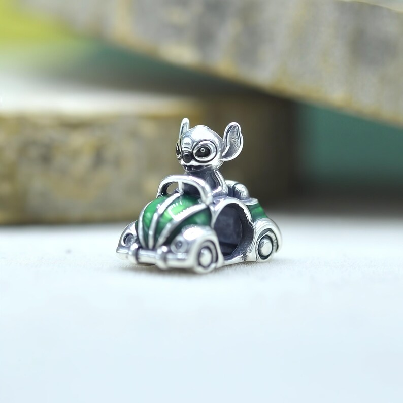 May include: A silver charm bead featuring the character Stitch driving a small green and silver car. The charm has a metallic finish with green enamel detailing on the hood and fenders, and a hollow center for a bracelet or necklace chain.