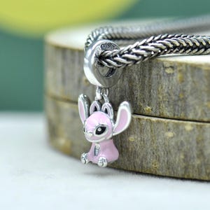 May include: A silver charm bracelet with a pink and silver Stitch charm. The charm features a sitting Stitch with large ears and a detailed face. The bracelet is made of woven silver links.