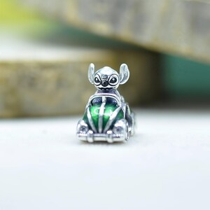 May include: A silver charm bead featuring the character Stitch from Lilo and Stitch driving a small green car. The character has large ears and black eyes, while the car has a glossy green finish with silver detailing on the headlights and bumper.