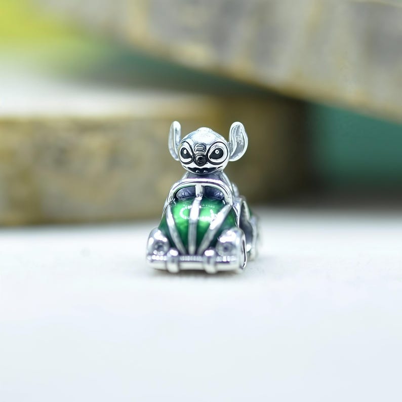 May include: A silver charm bead featuring the character Stitch from Lilo and Stitch driving a small green car. The character has large ears and black eyes, while the car has a glossy green finish with silver detailing on the headlights and bumper.