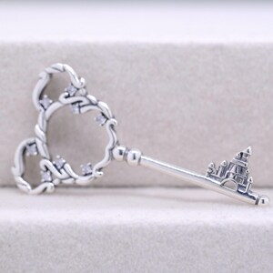 May include: A silver-colored key pendant with a decorative design. The key's head features a heart-shaped design with small, clear stones. The key's bit is shaped like a castle. A charming piece of jewelry.
