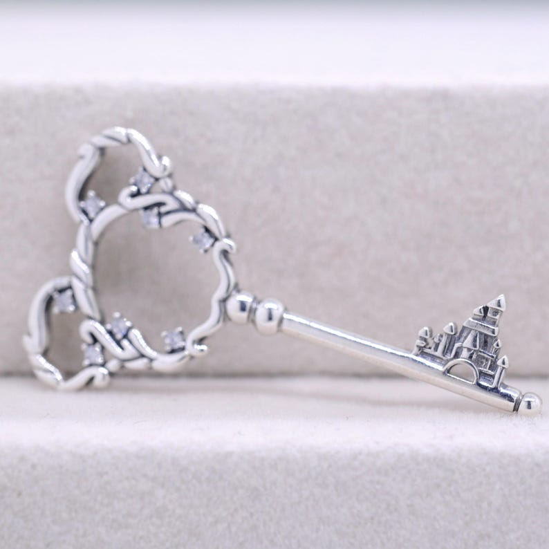 May include: A silver-colored key pendant with a decorative design. The key's head features a heart-shaped design with small, clear stones. The key's bit is shaped like a castle. A charming piece of jewelry.