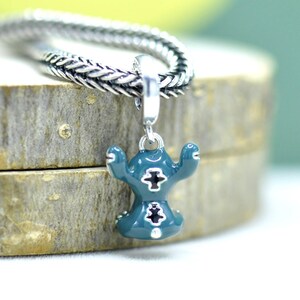 May include: A teal charm with a cartoon character design, featuring a silver cross detail. The charm is suspended from a silver chain. The charm is on a wooden surface.