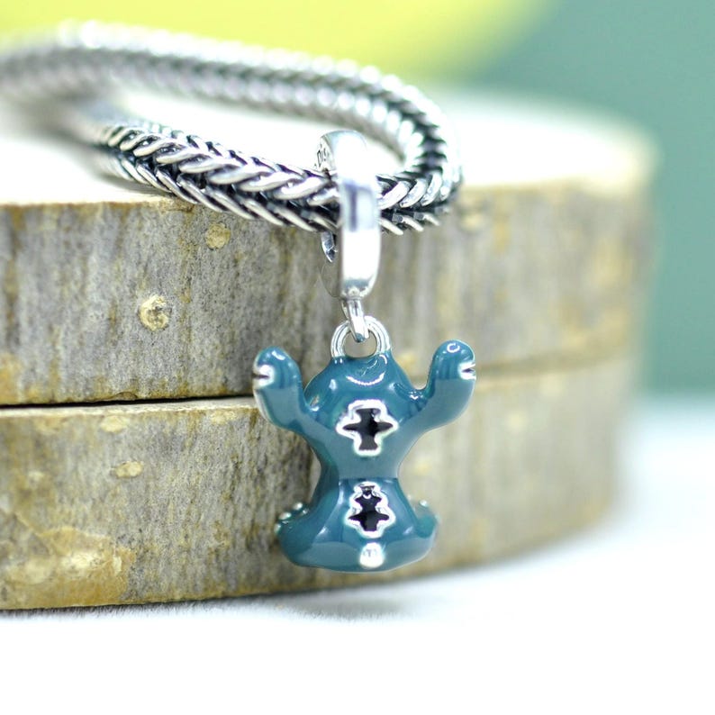 May include: A teal charm with a cartoon character design, featuring a silver cross detail. The charm is suspended from a silver chain. The charm is on a wooden surface.