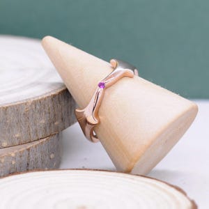 Può includere: Anello in oro rosa dal design unico e stilizzato. L'anello presenta una piccola gemma rosa rotonda ed è esposto su un porta anelli in legno. Il design dell'anello include linee curve e fluide ed elementi decorativi, mostrando un mix di eleganza e stile moderno.