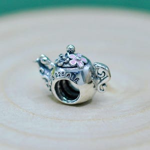 以下が含まれることがあります： ピンクの花のアクセントが付いた、銀色のティーポット型のチャーム。 チャームには、ハンドルと注ぎ口を含む複雑なディテールがあり、側面には「925 ALE」の文字が見えます。