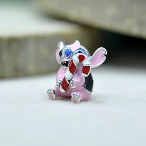 May include: A small metal charm shaped like a pink creature with large ears, blue accents on the head, and a wide grin, holding a red and white striped candy cane. The charm features enamel detailing and a silver-toned finish throughout the design.