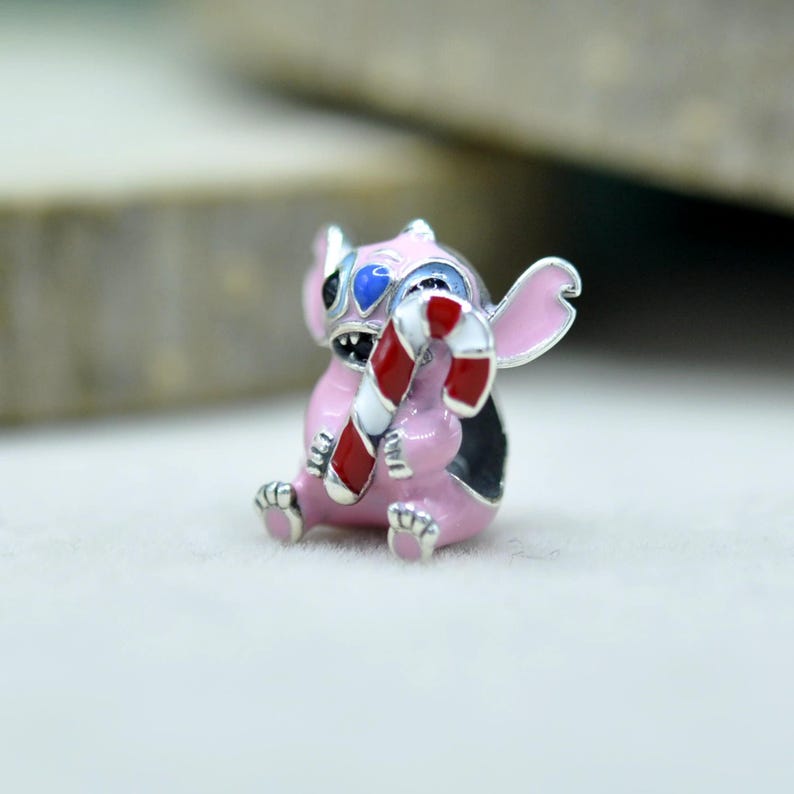 May include: A small metal charm shaped like a pink creature with large ears, blue accents on the head, and a wide grin, holding a red and white striped candy cane. The charm features enamel detailing and a silver-toned finish throughout the design.
