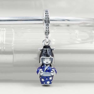 May include: A silver charm with a Japanese doll design. The doll is dressed in a blue kimono with white star patterns and a black hat. The charm has a silver loop with small, clear stones.