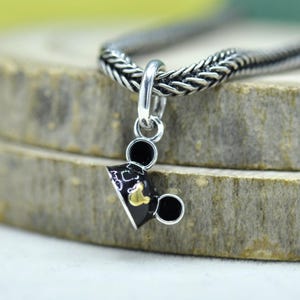May include: A silver charm with a black and gold Mickey Mouse graduation cap design. The charm is attached to a silver chain. The charm has two black circles on each side of the cap, and a gold emblem on the front.