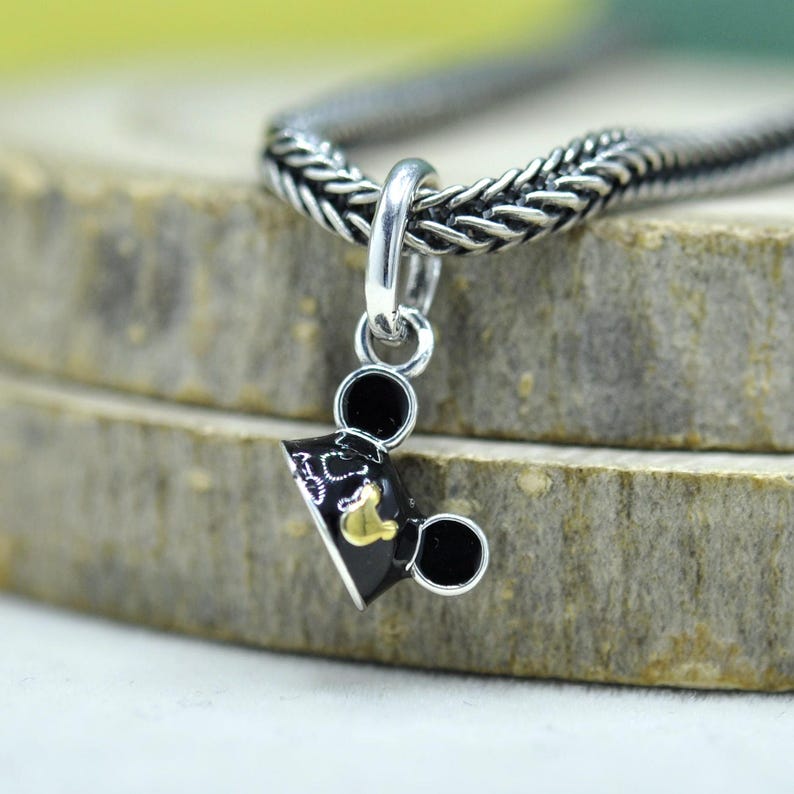 May include: A silver charm with a black and gold Mickey Mouse graduation cap design. The charm is attached to a silver chain. The charm has two black circles on each side of the cap, and a gold emblem on the front.