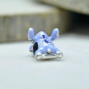 May include: A light blue enamel charm shaped like a cartoon creature with large ears and dark blue accents on the chest and head. The charm features a metallic silver base and a central hole for threading onto a bracelet or necklace chain.
