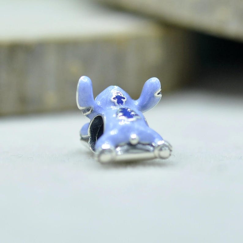 May include: A light blue enamel charm shaped like a cartoon creature with large ears and dark blue accents on the chest and head. The charm features a metallic silver base and a central hole for threading onto a bracelet or necklace chain.