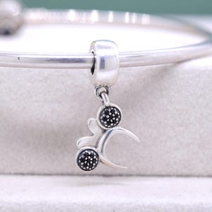 May include: A silver charm with a decorative design, featuring two circular elements with black accents and a curved shape. The charm is suspended from a silver bracelet.
