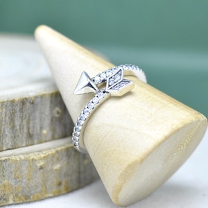 Könnte beinhalten: Ein silberner Pfeilring mit einem geometrischen Pfeildesign. Das Ringband ist mit kleinen, klaren Edelsteinen besetzt. Pfeilspitze und Befiederung sind mit einer polierten Oberfläche versehen. Der Ring wird auf einem Holzringhalter präsentiert.