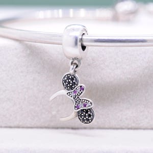 May include: A silver charm with a Minnie Mouse design. The charm features a round bead with black stones, a bow with purple and clear stones, and a crescent shape. The charm is attached to a silver bracelet.