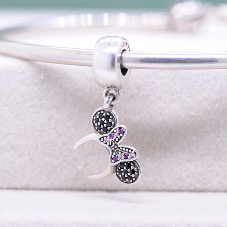 May include: A silver charm with a Minnie Mouse design. The charm features a round bead with black stones, a bow with purple and clear stones, and a crescent shape. The charm is attached to a silver bracelet.
