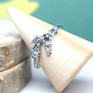 May include: A silver ring with a detailed floral and star design. The ring is displayed on a wooden stand, showcasing its intricate craftsmanship. The ring's design includes small, sparkling accents, adding to its delicate appearance.