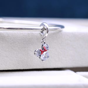 May include: Silver charm with a red bow and clear crystals, shaped like Minnie Mouse. The charm is attached to a silver bracelet.