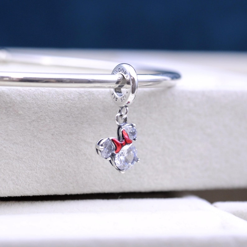 May include: Silver charm with a red bow and clear crystals, shaped like Minnie Mouse. The charm is attached to a silver bracelet.