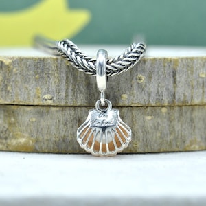 Może przedstawiać: Srebrny charms w kształcie muszli z wygrawerowanym napisem "One of a kind". Charms jest przymocowany do srebrnego łańcuszka. Muszla ma jasno pomarańczowe wnętrze i jest umieszczona na drewnianym tle.