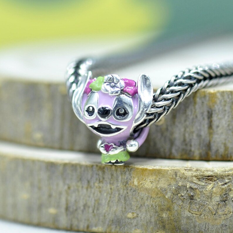 May include: A silver charm bead featuring a light purple character with large ears, a pink flower crown, and a green skirt, threaded onto a silver chain. The character has black eyes, a black nose, and a small pink heart held in its hands.