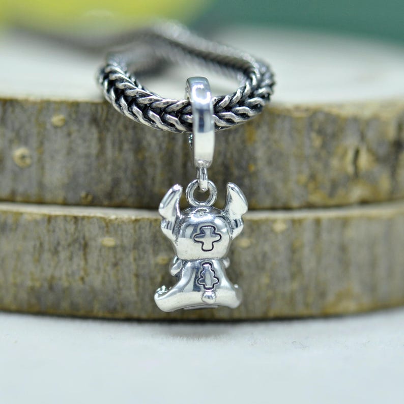 May include: A silver charm pendant featuring a stylized character with large ears and a cross symbol on its chest. The charm is suspended from a silver chain, with a braided texture. The pendant is set against a wooden background.