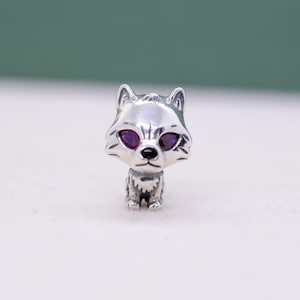 Może przedstawiać: Srebrny charms w kształcie wilka, z fioletowymi oczami z kamieni szlachetnych. Wilk jest przedstawiony w pozycji siedzącej, ze szczegółami sierści i rysów twarzy. Ten charms jest prawdopodobnie do bransoletki lub naszyjnika.