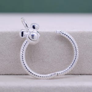 Pu&ograve; includere: Orecchino a cerchio color argento con un design ispirato a Mickey Mouse. L'orecchino presenta un cuore al centro e una fascia a spirale strutturata. Il design include due piccole forme rotonde e una piccola forma curva.