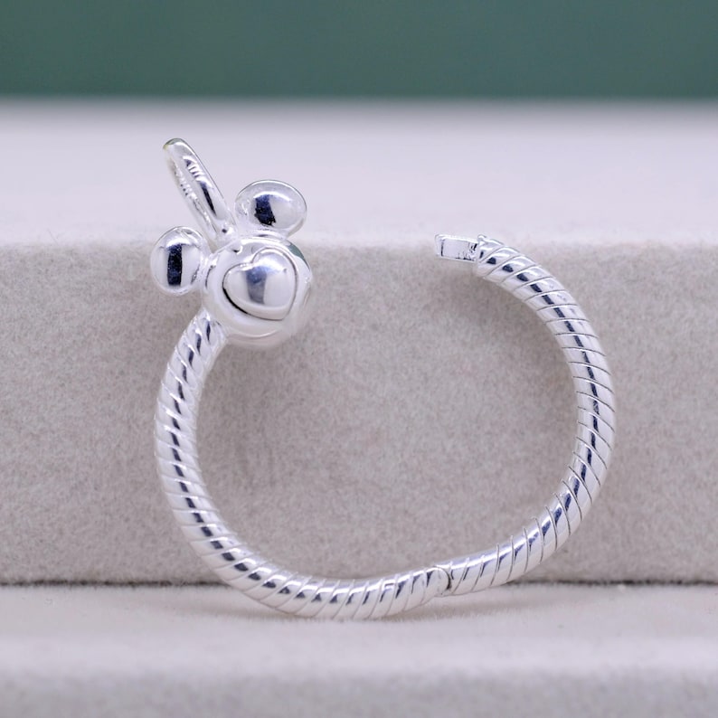 Pu&ograve; includere: Orecchino a cerchio color argento con un design ispirato a Mickey Mouse. L'orecchino presenta un cuore al centro e una fascia a spirale strutturata. Il design include due piccole forme rotonde e una piccola forma curva.