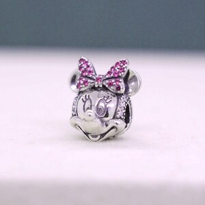 May include: A silver charm featuring a Minnie Mouse design. The charm has a bow with pink gemstones and white gemstones around the face. The charm has two round ears and a smiling face.