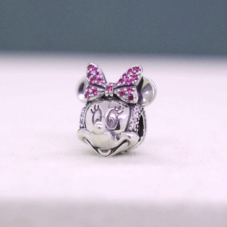 May include: A silver charm featuring a Minnie Mouse design. The charm has a bow with pink gemstones and white gemstones around the face. The charm has two round ears and a smiling face.