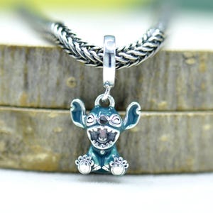 May include: A teal and silver charm featuring the character Stitch from the animated film Lilo & Stitch. The charm is detailed with black and pink accents, and hangs from a silver chain. The charm is a pendant.