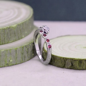 May include: A silver ring with a dragon design. The ring features a dragon head and a curved band adorned with small, red gemstones. The ring is set against a light background with green wooden slices.