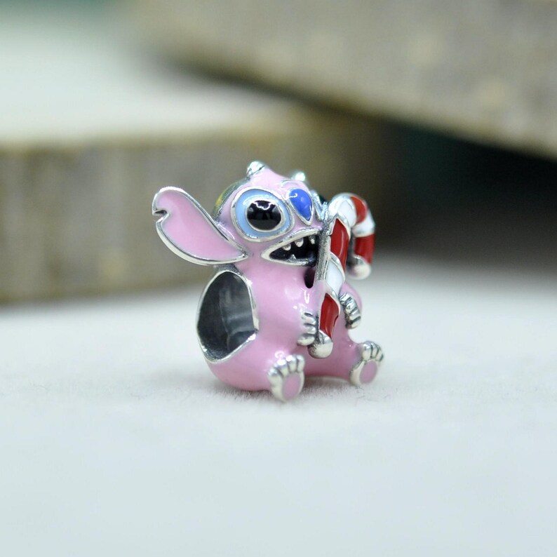 May include: A silver charm bead shaped like a pink creature with large ears, blue eyes, and a wide grin, holding a red and white striped candy cane. The charm features enamel detailing in pink, blue, red, and white, with a hollow center for a bracelet.