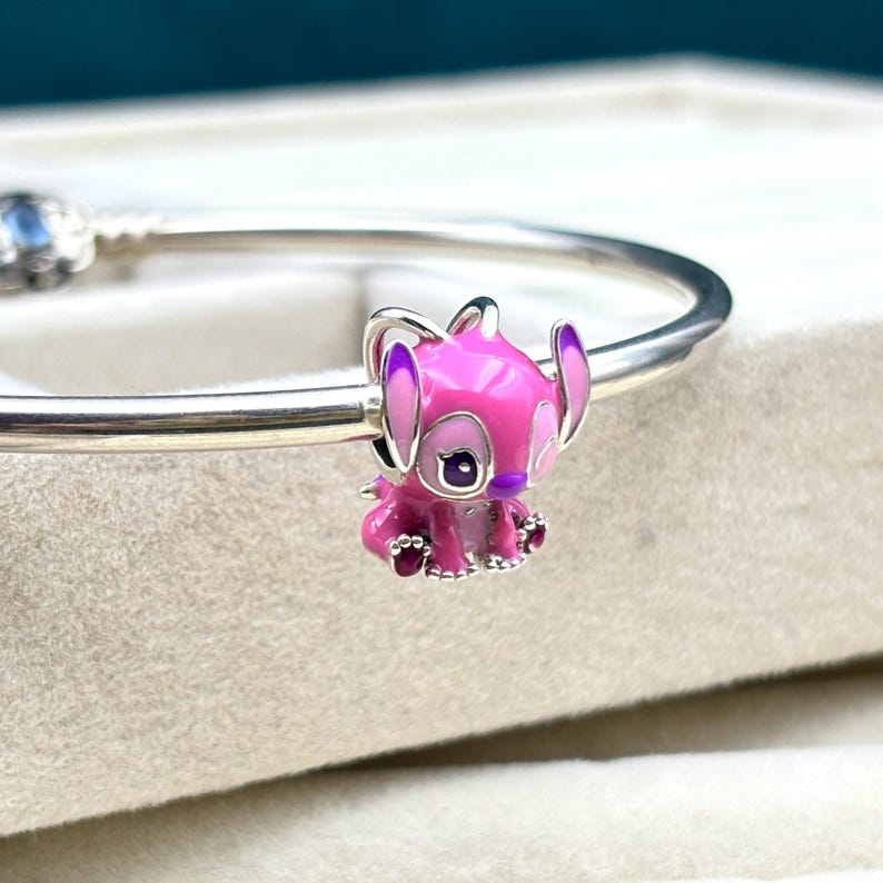 May include: A pink and purple enamel charm of Stitch, a character from the Disney movie Lilo & Stitch, sitting on a silver bracelet.