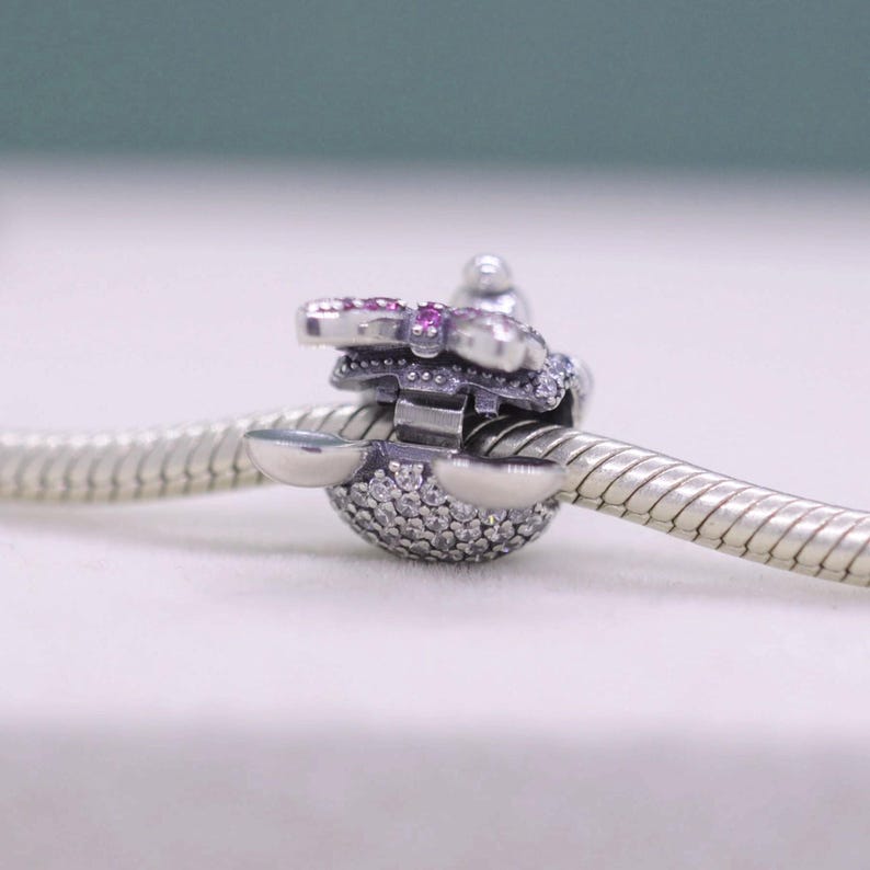 May include: A silver charm with a butterfly design, featuring pink and clear gemstones. The charm is attached to a silver snake chain bracelet. The butterfly has pink gemstones on the wings and clear gemstones on the body.