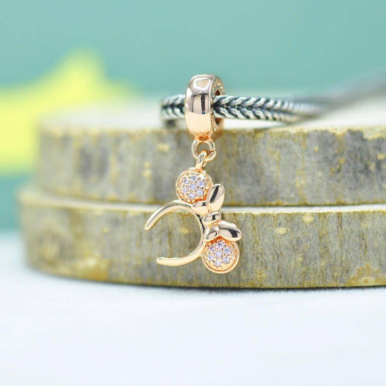 May include: A gold-colored charm with a Mickey Mouse-inspired design. The charm features a bow and two circular elements, all adorned with small, sparkling crystals. The charm is attached to a silver-colored braided ring.