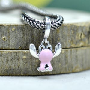 May include: A silver charm pendant featuring a pink cartoon character with outstretched arms. The charm is attached to a silver chain. The pendant is set against a wooden background.